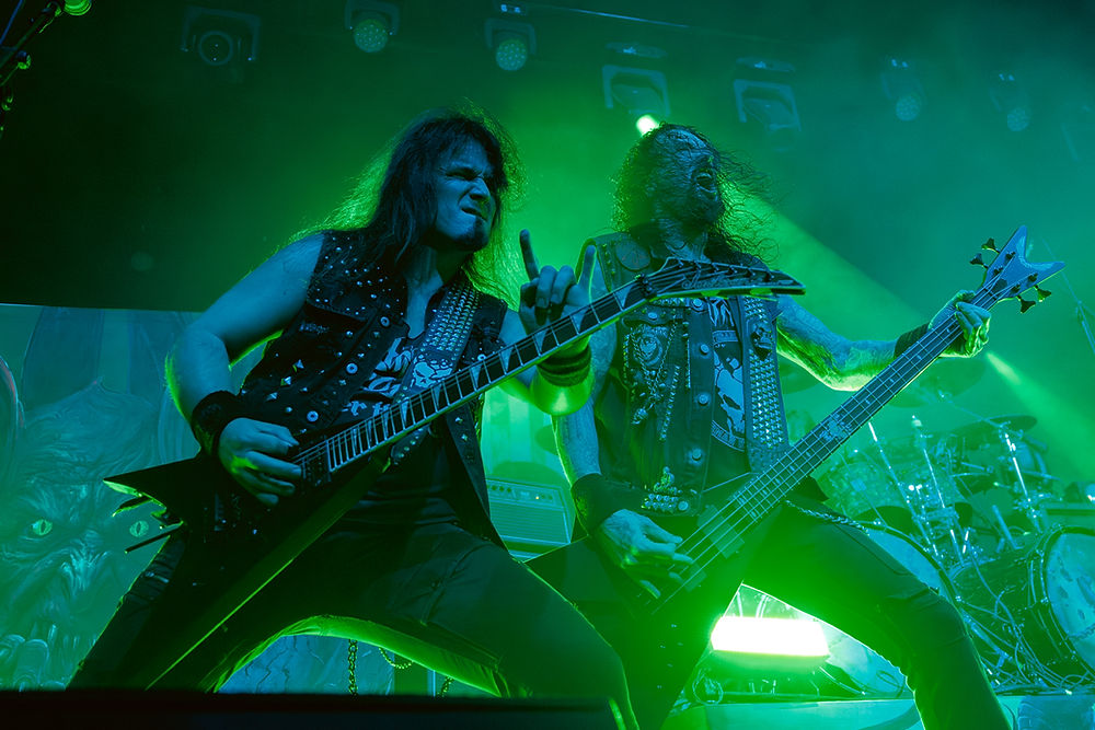 Rock musicians perform intensely on stage with electric guitars. They're bathed in green light, wearing studded vests. Energetic atmosphere.