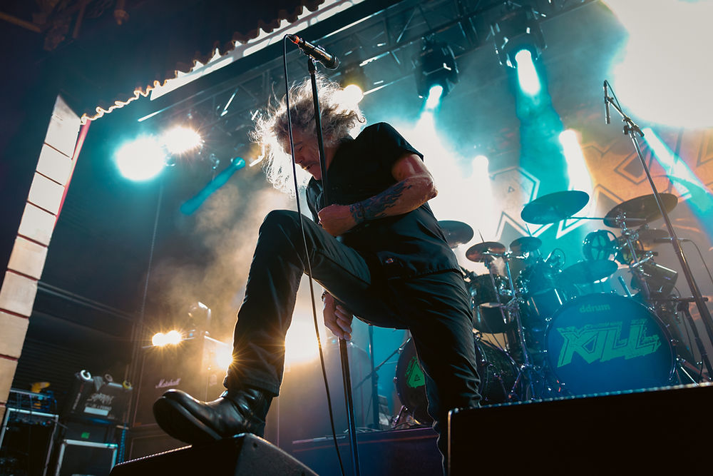 Singer passionately performing on stage, holding a microphone stand. Bright stage lights and drum set in the background create a dynamic atmosphere.