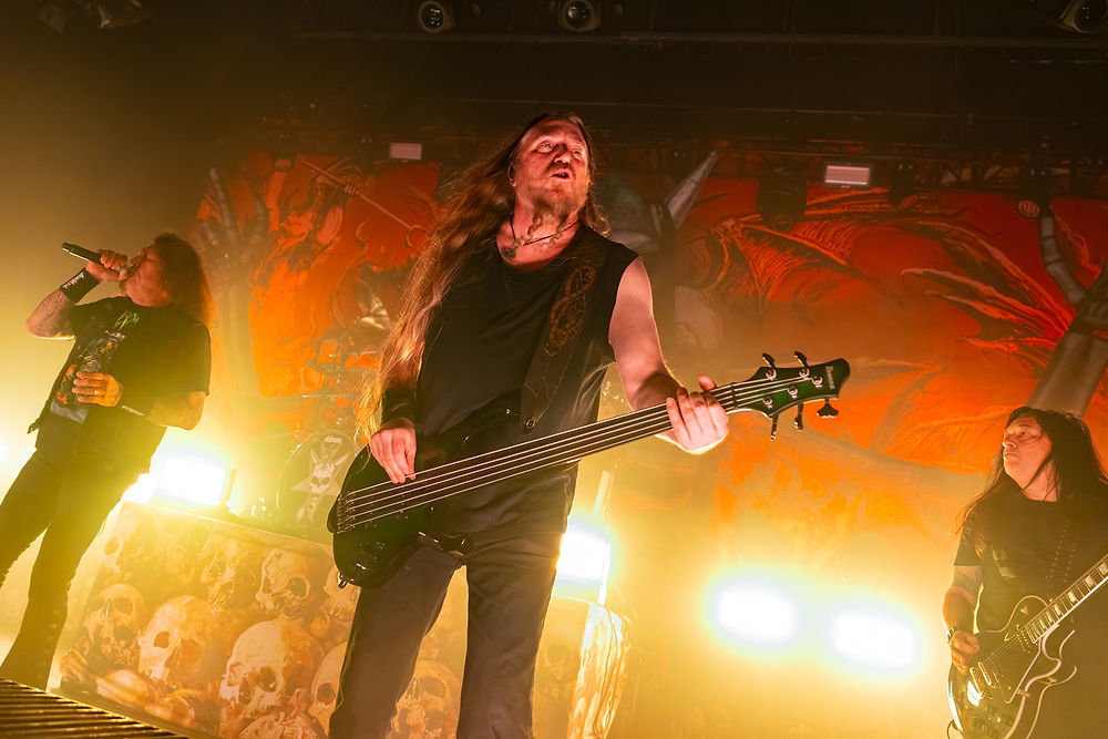 Band performs energetically on stage with vivid orange backdrop. Vocalist sings with mic, bassist plays guitar, intense stage lighting.
