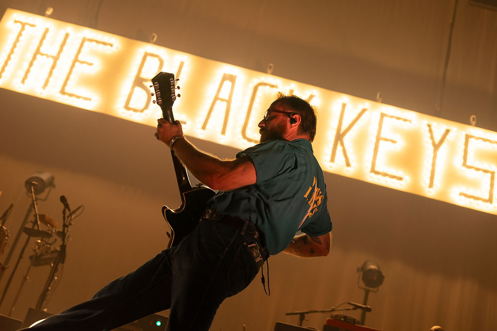 Guitarist on stage with a tilted body, vibrant lighting, and "THE BLACK KEYS" sign in the background. Energetic, dynamic atmosphere.
