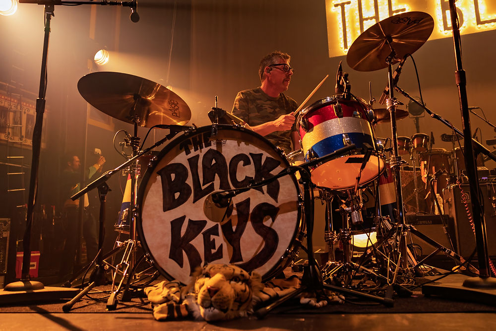 Drummer performs energetically on a stage with "The Black Keys" drum kit. Dim lighting, colorful drum, and toy tiger in foreground.