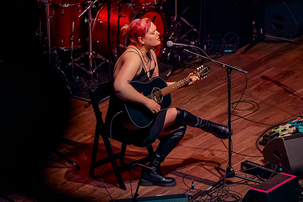 Musician with pink hair playing guitar and singing onstage. She sits in a wooden chair, surrounded by drums and cables, wearing black boots.