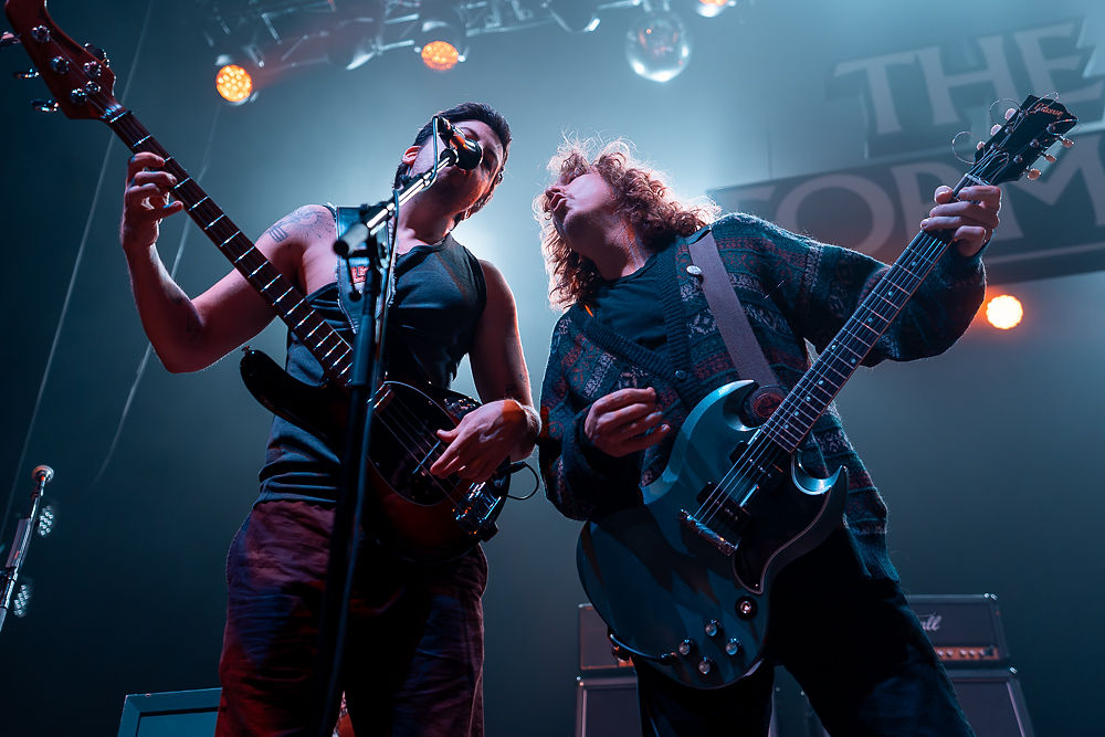 Two musicians perform on stage under blue and orange lights, playing electric guitars. A backdrop reads "THE". Energetic atmosphere.
