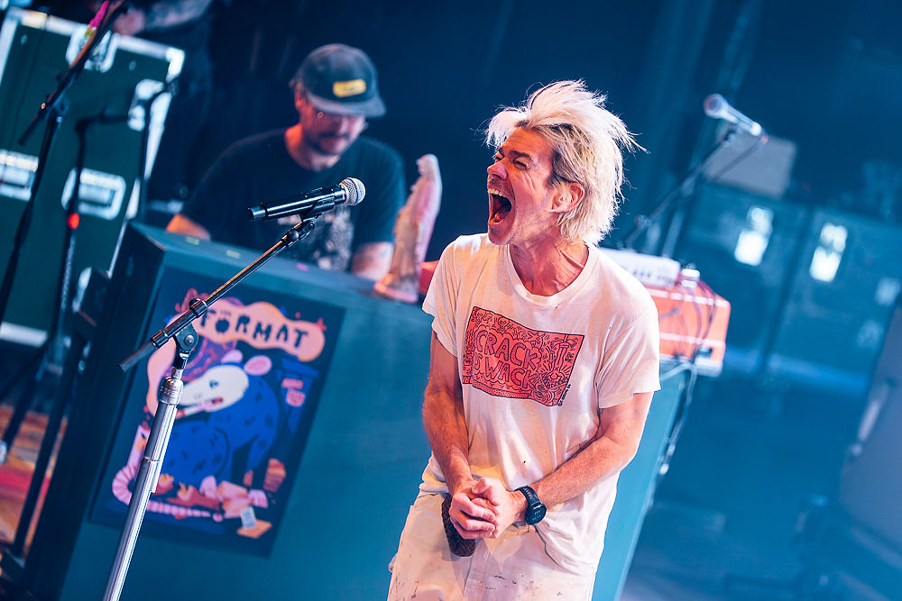 Singer passionately performing on stage, wearing a white "Crack Wack" shirt. Vibrant blue poster and keyboard in the background.