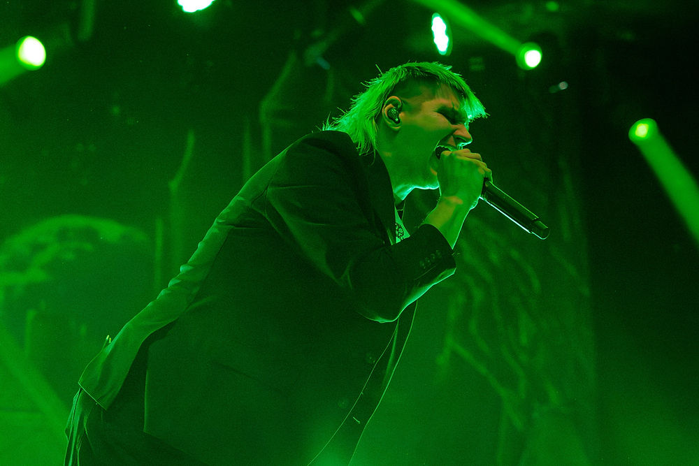 Singer passionately performs on stage under green lights, holding a microphone. Background shows abstract patterns and more green lighting.