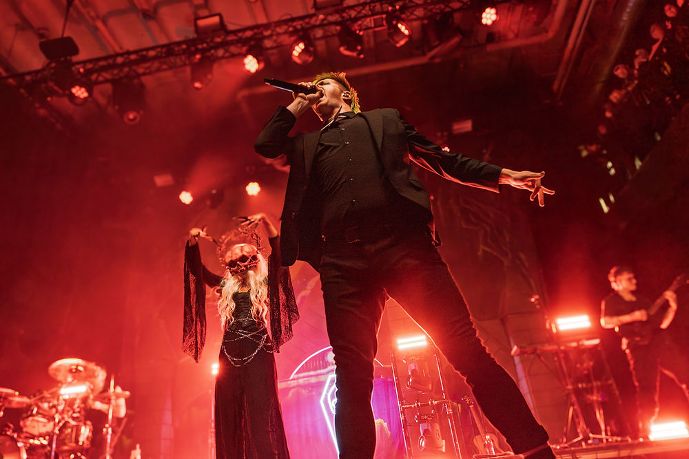 Singer on stage with mic, red lighting; person in mask and crown poses dramatically. Concert ambiance with drums and guitar in background.