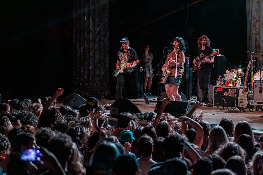Band performing on stage with guitars and a bass. Crowded audience cheers under dim lighting. Energetic atmosphere, stage decorated with props.