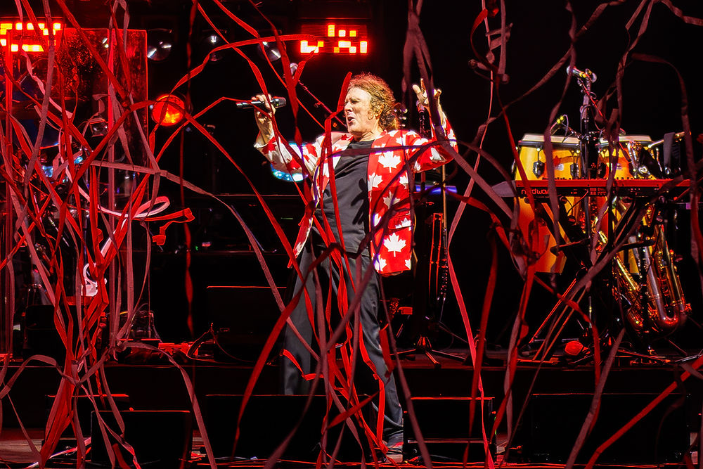 Singer in a red maple-leaf jacket performs on a dark stage, holding a mic amid red streamers and glowing lights.
