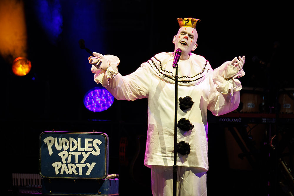Pale clown in white costume sings at a microphone on a dark stage, beside a suitcase reading Puddles Pity Party.