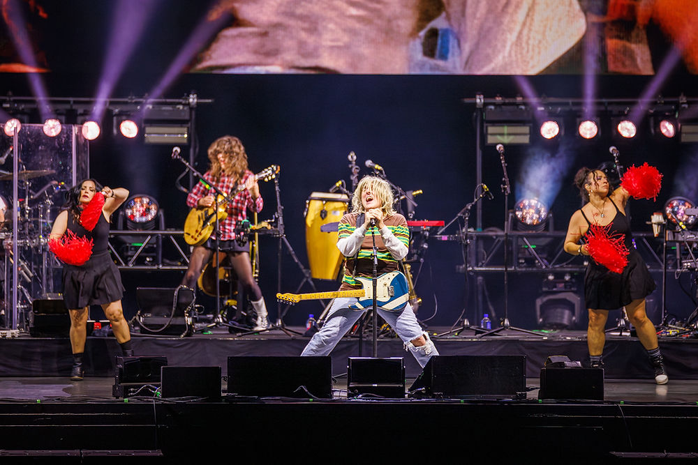 Rock band performing on a lit stage, singer at center with guitar, dancers waving red pom-poms, dramatic concert energy