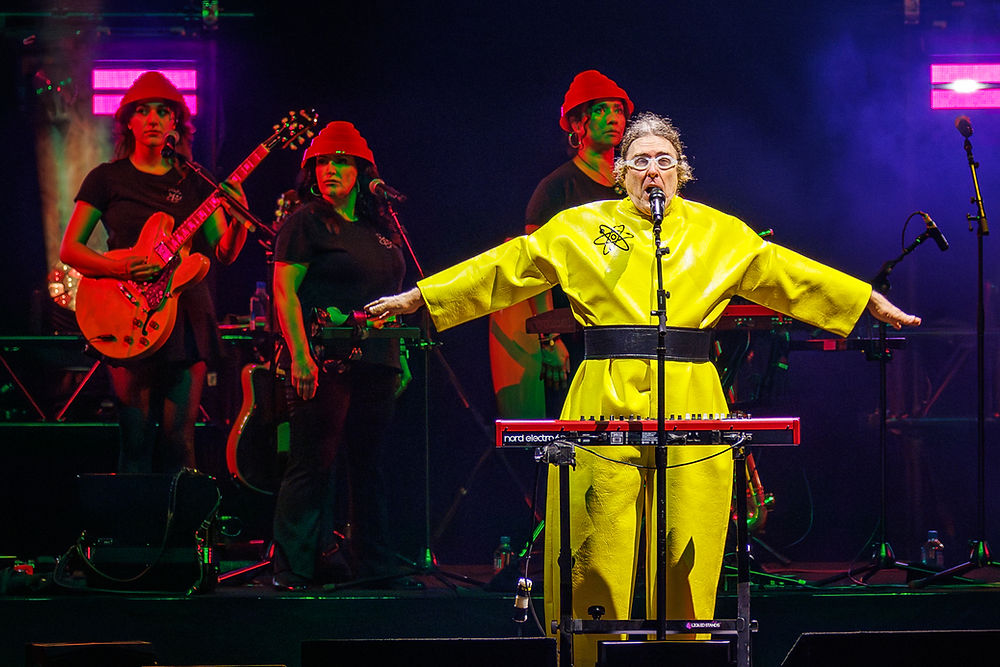 Singer in bright yellow stands at a mic with arms out on a neon-lit stage, backed by guitarists in red hats.