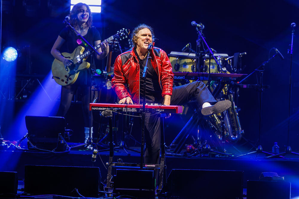 Singer in red jacket plays keyboard and sings on blue-lit stage, with guitarist and drums behind; nord electro visible.