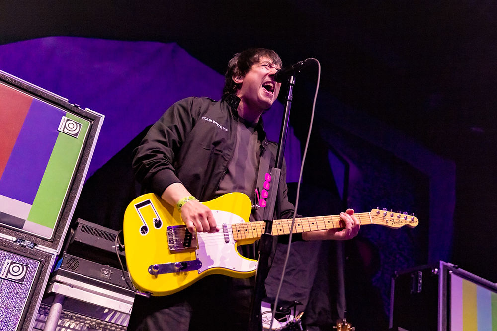 Musician passionately plays a yellow guitar on stage, singing into a microphone. Vibrant backdrop with colorful amplifiers, energetic mood.