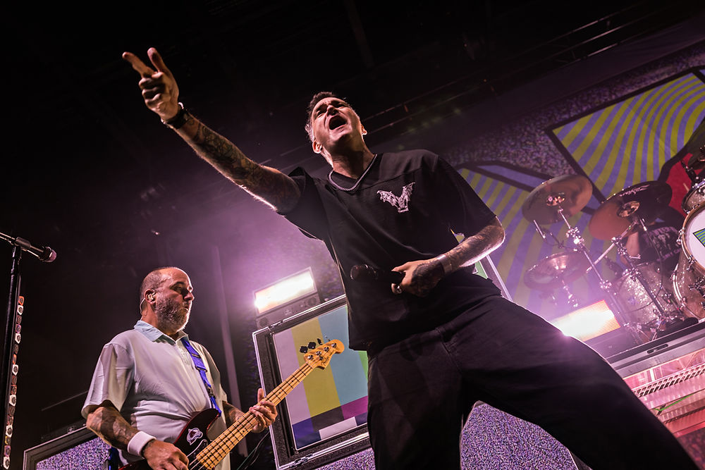 Rock band performing on stage with colorful geometric background. Singer gestures energetically while guitarist plays. Dynamic and vibrant scene.