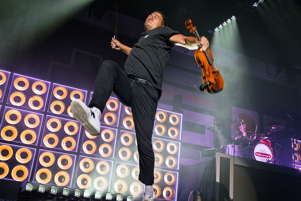 Musician jumping with a violin on stage, near speaker lights. Drummer in the background. Energetic performance with dynamic lighting.