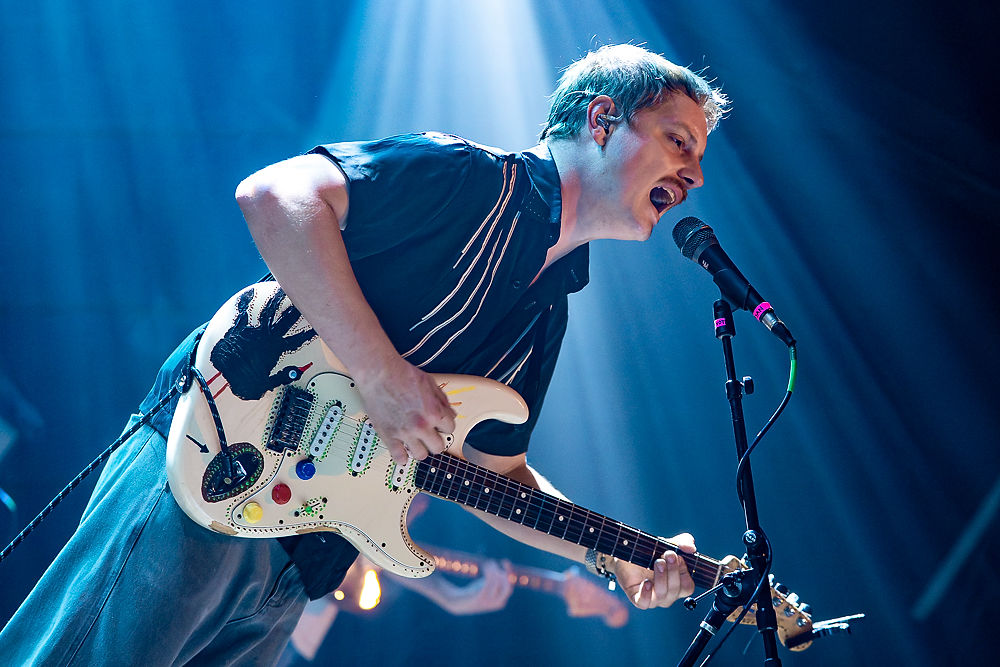 Musician passionately sings into a microphone on stage, playing a decorated electric guitar under blue spotlights. Energetic performance.