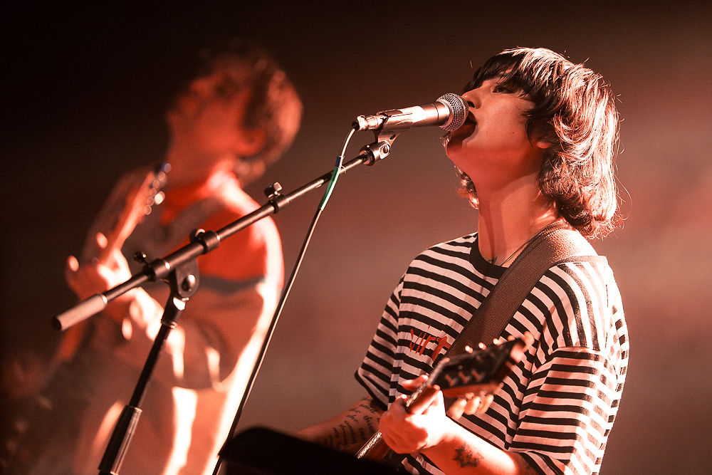 Musician in striped shirt sings into a microphone while playing guitar. Blurred guitarist in the background. Warm stage lighting.