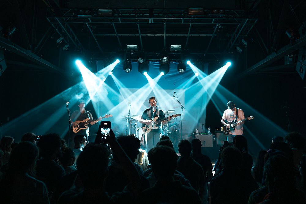 Band performing on stage with blue spotlights, surrounded by a crowd. Musicians play guitars and drums, creating lively atmosphere in dark venue.