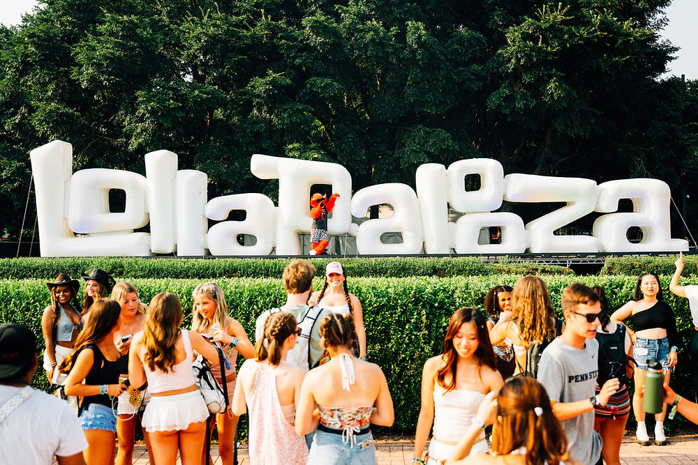 Crowd of people smiling in front of large inflatable "Lollapalooza" sign. Bright day, trees in background, festive atmosphere.