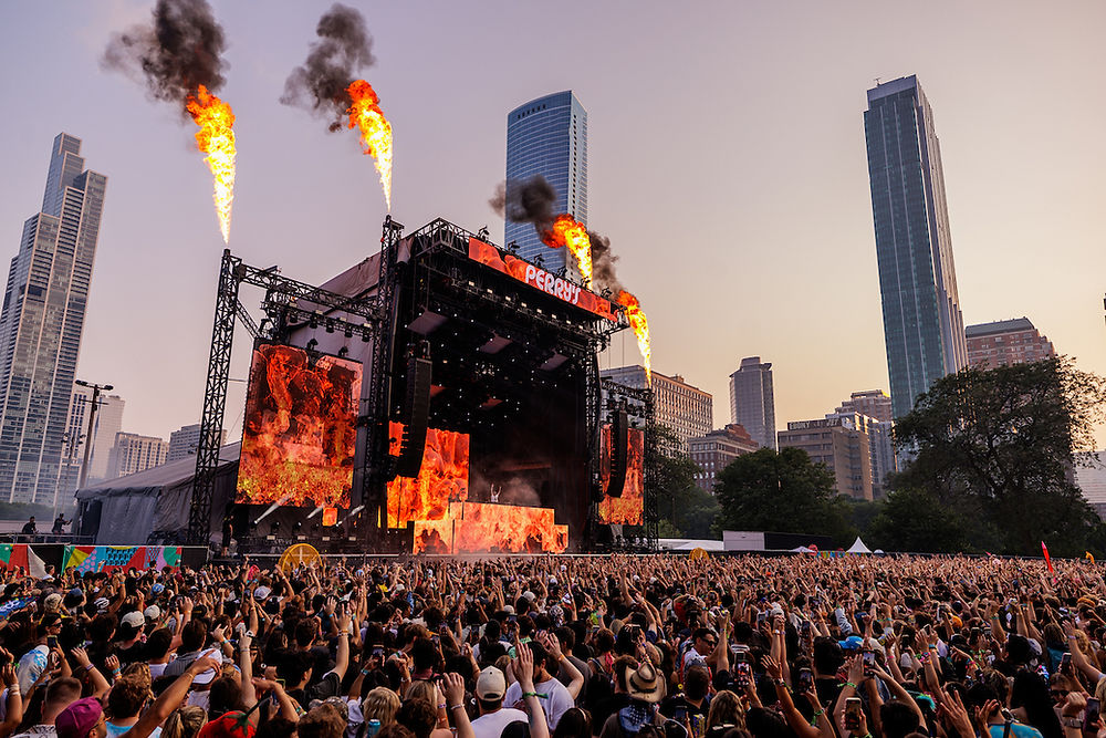Crowd watches a fiery stage performance at sunset in a city park, with flames erupting above. Skyscrapers tower in the background.
