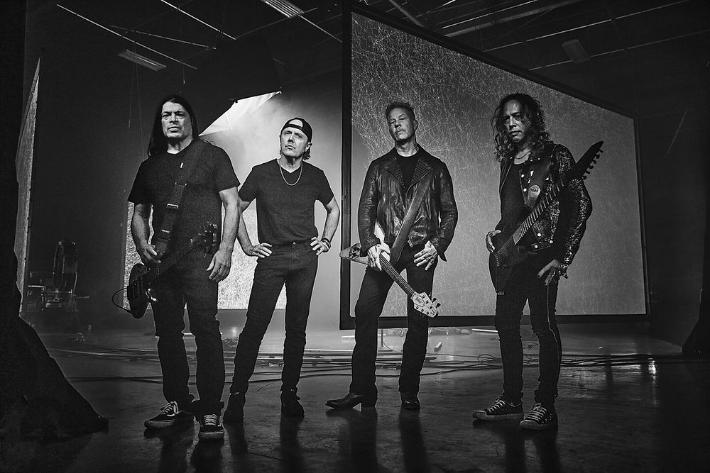 Four men with guitars stand confidently in a dimly lit studio. Black outfits and intense expressions create a dramatic rock band vibe.