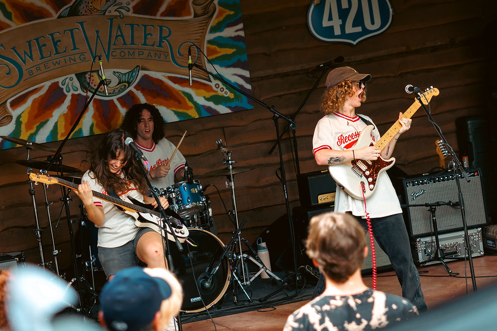 Band performing live on stage with colorful backdrop and SweetWater Brewing sign. Guitarist and bassist in jerseys, audience in foreground.