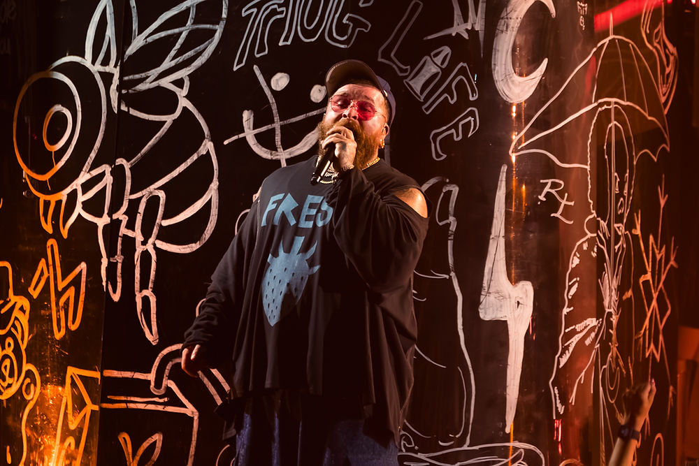 Singer in a cap and pink glasses performs with a mic. Black shirt reads "FRESH." Graffiti backdrop with white drawings and text. Energetic vibe.