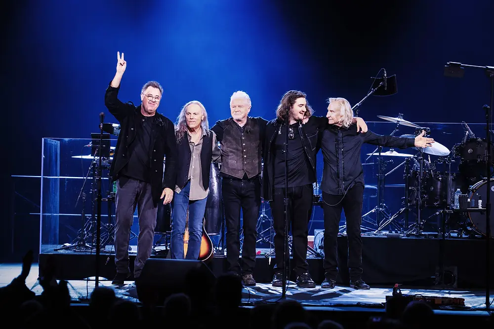 Five men stand on stage, smiling, arms around each other. One holds a peace sign. Drums in the background. Blue lighting, live concert.
