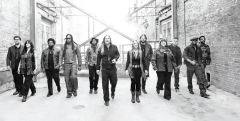 Group of people walking in an urban alley with brick walls, wearing casual and patterned clothing. The mood is relaxed and confident.