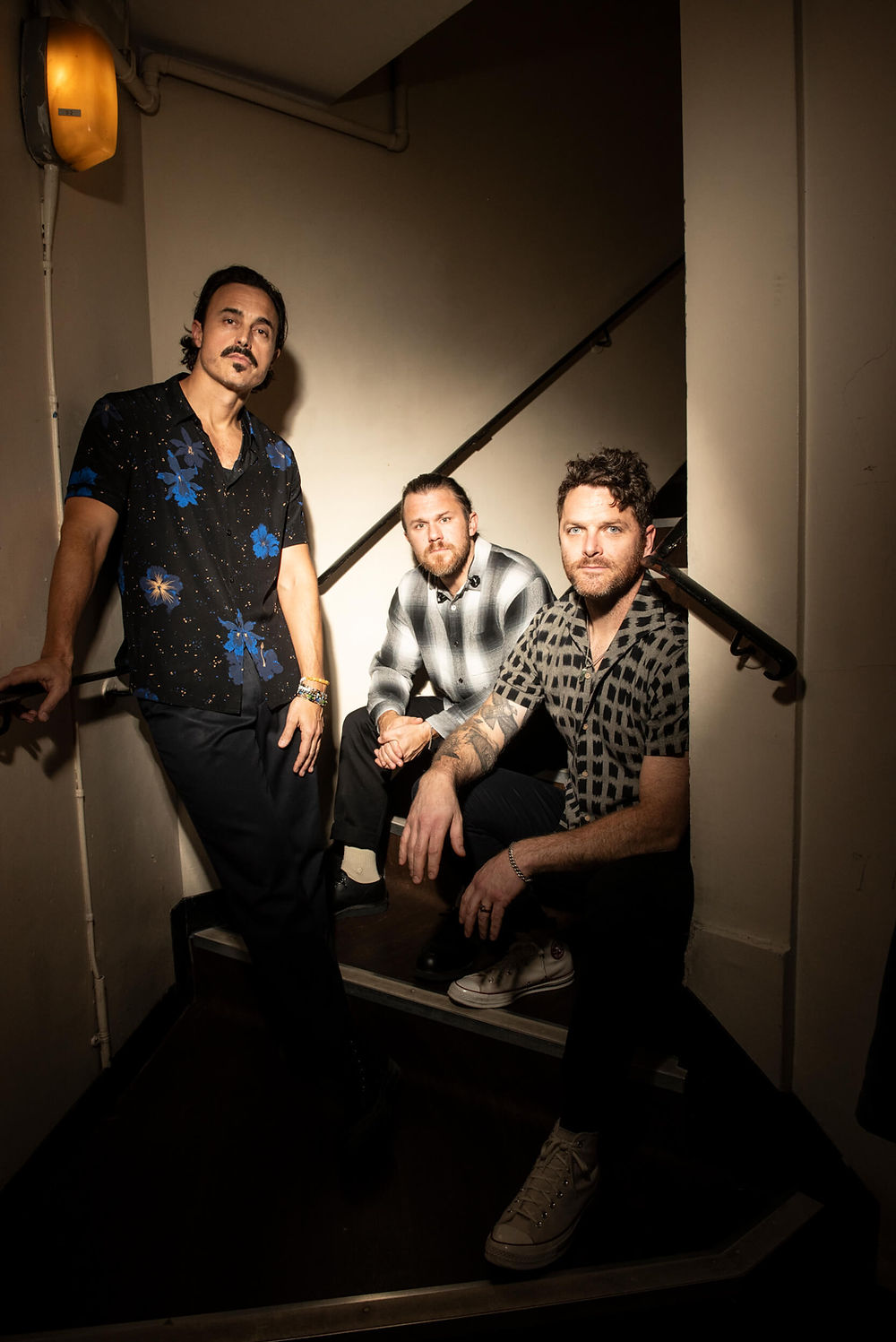 Three men pose on dimly lit stairs in stylish shirts, creating a moody atmosphere. One stands, while two crouch, highlighting varied patterns.