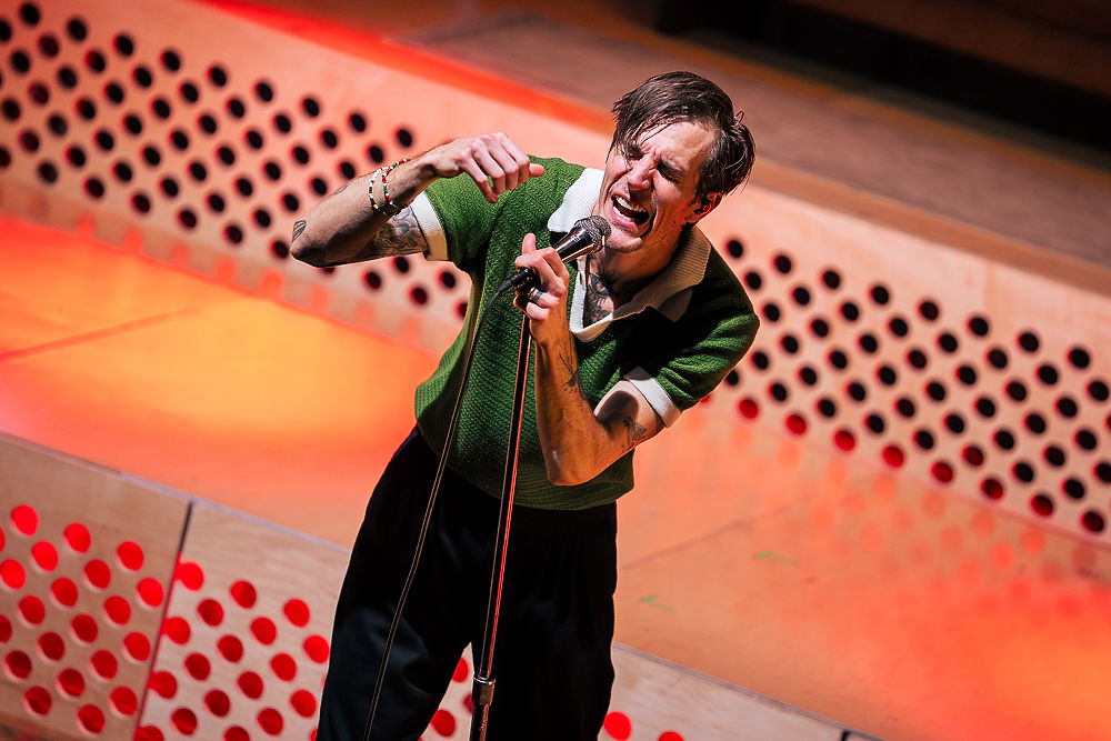 Singer in a green shirt passionately performs onstage, holding a microphone. The background has orange lighting with circular patterns.