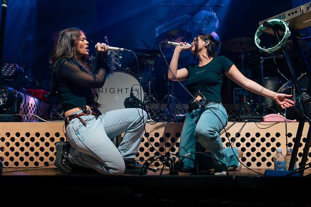 Two women sing passionately on stage, kneeling with microphones. Blue and black stage lights and instruments set the energetic concert scene.