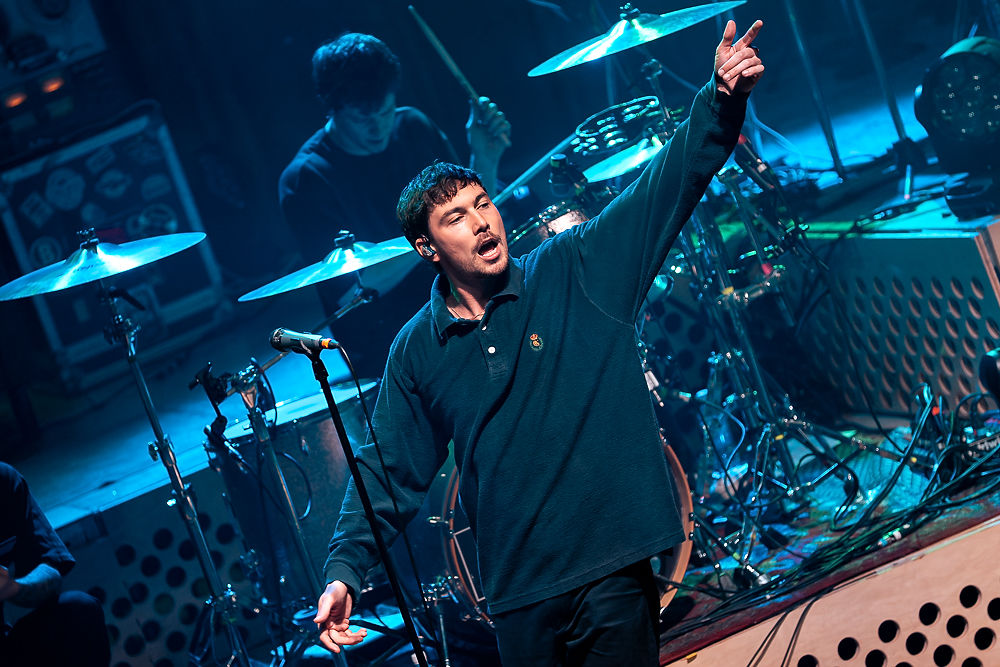 Singer passionately performs on stage, arm raised, with a drummer in the background. Blue lighting creates a vibrant concert atmosphere.
