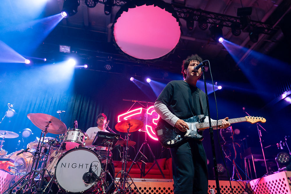 Musician sings and plays guitar on stage, with a drummer nearby. Bright stage lights and a neon sign create a lively concert atmosphere.