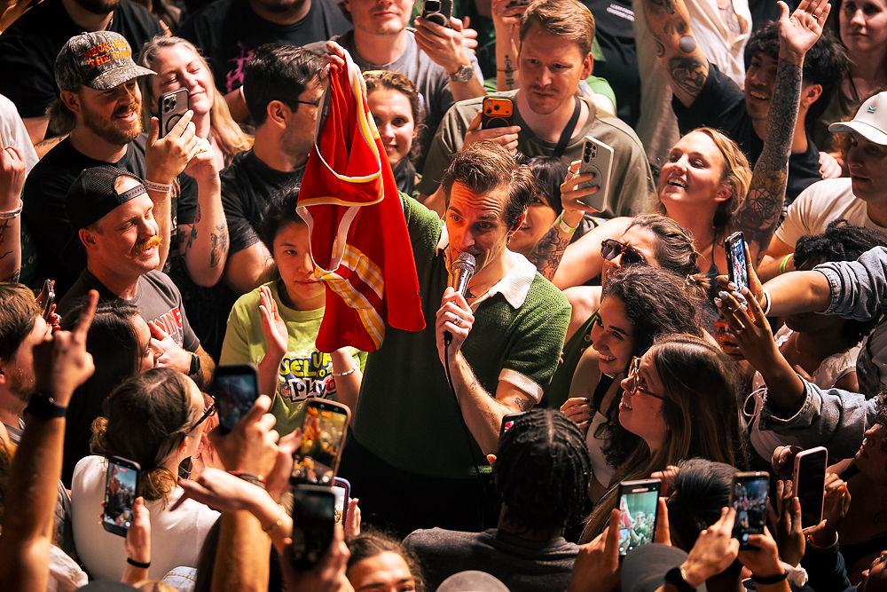 Man in green shirt holds red jersey, singing into mic, surrounded by smiling crowd with phones. Energetic concert atmosphere.