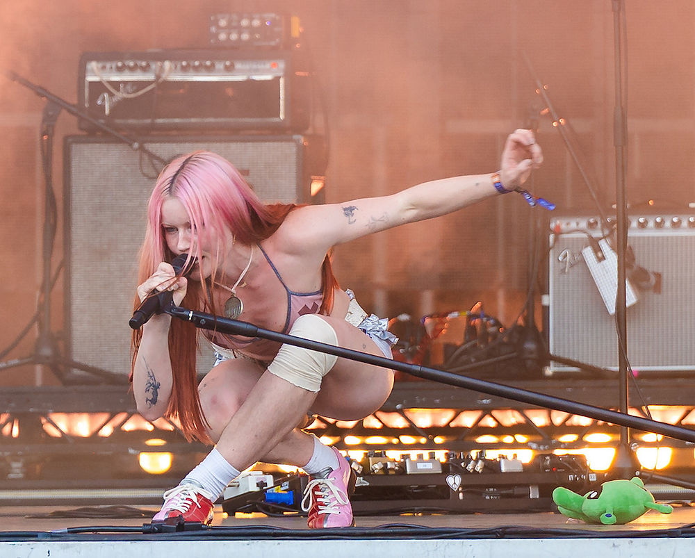 A person with pink hair sings energetically into a mic on stage. They wear casual attire with knee pads. Background features amplifiers.