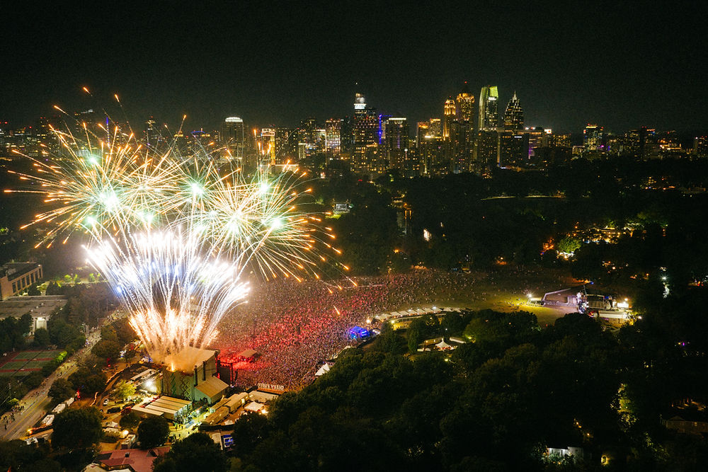 Fireworks burst over a crowded outdoor concert at night, with a vibrant city skyline in the background. The mood is festive and lively.